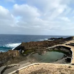 Piscinas naturales formadas por lava volcánica en la costa de Lanzarote, con el mar azul de fondo. No hay cocoteros, pero sí un paisaje salvaje.
