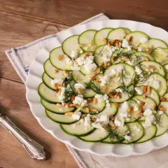 Delicioso carpaccio de calabacín con queso feta desmenuzado, piñones tostados y eneldo fresco, servido en un plato blanco sobre una mesa de madera.