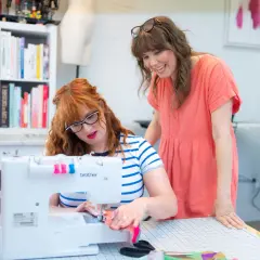 Two women are focused on sewing seams on a white Brother sewing machine in a bright studio.