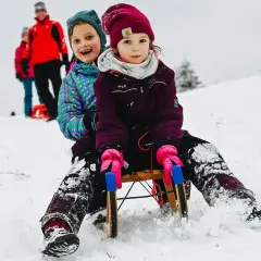 Zwei Kinder rodeln lachend einen verschneiten Hang im Bergischen Land hinunter.