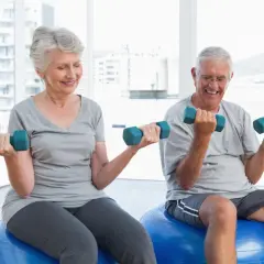Uśmiechnięta para seniorów ćwiczy z hantlami na piłkach gimnastycznych, dbając o zdrowie i zapobiegając osteopenii.