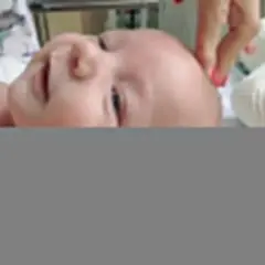 Uroczy niemowlak z delikatnymi zakolami, leżący na białym tle. Dłoń z pomalowanymi paznokciami delikatnie dotyka jego główki.