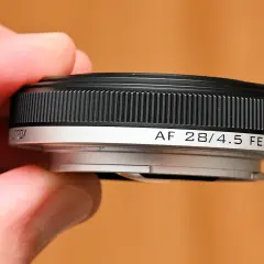 A hand holds a compact, black and silver pancake lens, labeled "AF 28/4.5 FE".
