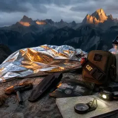 Equipo de bushcraft que es: manta térmica, cuchillo, pedernal, brújula, mapa, botiquín, cantimplora y frontal, listos para la aventura en la montaña.