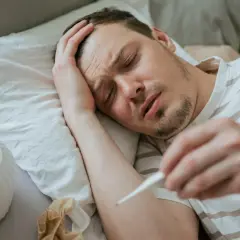 Chory mężczyzna leży w łóżku, trzymając termometr. Widać jego cierpiącą twarz i rękę na czole. To typowe objawy grypy wariant k.