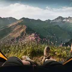 Vista desde una tienda de campaña: piernas con botas de montaña, flores silvestres y montañas verdes bajo un cielo nublado. Disfrutando del acampar con tienda de campaña.