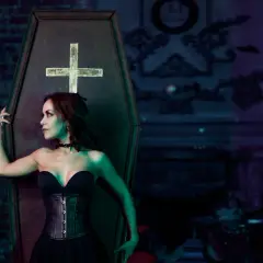 Kobieta w czarnym gorsecie i sukni opiera się o trumnę z krzyżem.