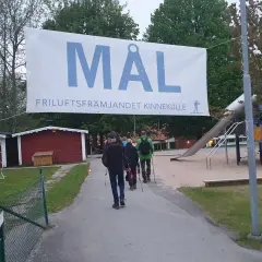A banner reading "MÅL FRILUFTSFRÄMJANDET KINNEKULLE" hangs over a path where people walk towards a playground.