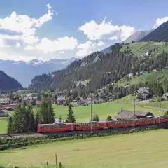 Ein roter Zug fährt durch eine malerische Alpenlandschaft. Ein perfekter tagesausflug mit der bahn, vorbei an grünen Wiesen, Wäldern und einem Dorf mit schneebedeckten Bergen im Hintergrund.
