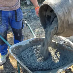 Robotnik w pomarańczowych rękawicach wylewa świeży beton z betoniarki do taczki. Różne rodzaje betonu wymagają różnych metod mieszania i transportu.