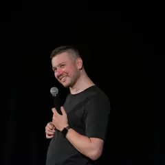 Grzegorz Wójtowicz w trakcie występu stand-up, trzyma mikrofon i uśmiecha się do publiczności.