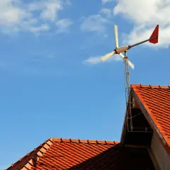 Mała przydomowa elektrownia wiatrowa na tle błękitnego nieba z chmurami, zamontowana na dachu.