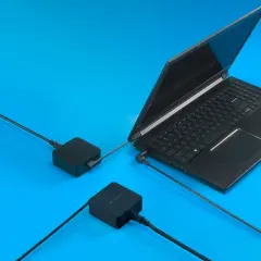 Czarny laptop gamingowy podłączony do dwóch zasilaczy na niebieskim tle.
