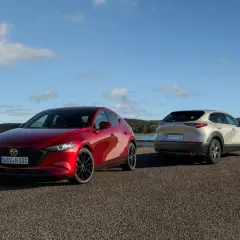 Dwie Mazdy: czerwona hatchback i beżowy SUV, stoją nad jeziorem. Sprawdź mazda cx 30 cena i wybierz swój wymarzony model.