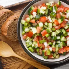 Ensalada pipirrana fresca y colorida, con tomate, pepino, pimiento y cebolla, servida junto a rebanadas de pan integral y una cuchara de madera.