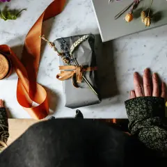Händer som förbereder sig för att slå in runda paket med grått papper, orange sidenband och torkade blommor.