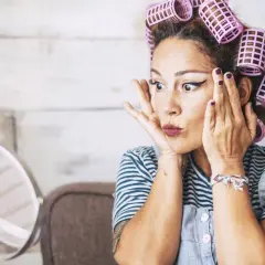 Mujer con rulos en el pelo, delineado de ojos y labios pintados, practicando cómo rizar el pelo frente a un espejo.