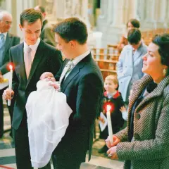 Un homme tient un bébé dans une robe de baptême blanche, une bougie allumée à la main. Il est le parrain de la famille, entouré d'autres membres de la famille dans une église.