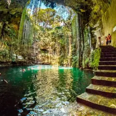 Vista interior del cenote Chichén Itzá, con escaleras de piedra y nadadores en sus aguas turquesas, rodeado de vegetación colgante.