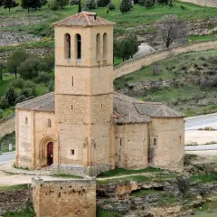 La iglesia de la Veracruz, de piedra y con un campanario robusto, se alza sobre un paisaje verde y ondulado, con un camino serpenteante a su lado.