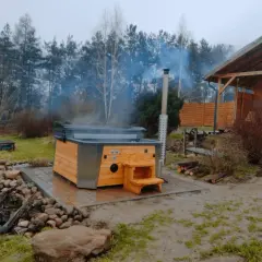 Drewniana balia z parującą wodą, obok której stoi domek w lesie. Idealne miejsce na relaks.
