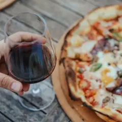 Un bicchiere di vino rosso in mano, accanto a una gustosa pizza. Un abbinamento perfetto per una serata all'aperto.