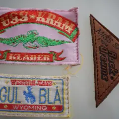 Tre märken: ett rosa med texten "PUSS & KRAM JEANS" och en grön drake, ett brunt triangelformat med "MISS SALLY FROM Rock Dreams", och ett vitt med "WESTERN JEANS GUL BLÅ WYOMING".