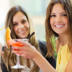 Dwie uśmiechnięte kobiety z drinkami. Jedna trzyma biały koktajl, druga pomarańczowy. Czy wódka tuczy?