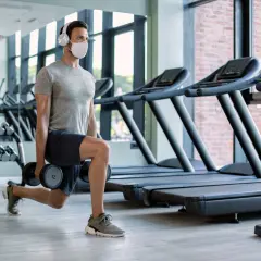 Mężczyzna w masce i słuchawkach wykonuje wykroki z hantlami. To jego trening cardio na siłowni, gdzie dba o formę.