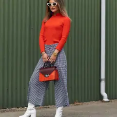 Kobieta w pomarańczowym golfie i kraciastych kulotach prezentuje stylowe kuloty stylizacje. Białe kozaki i torebka dodają charakteru.
