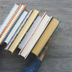 Ręce w dżinsowej kurtce trzymają stos kolorowych książek. To idealne nowości warte przeczytania na jesienne wieczory.