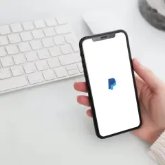 Dłoń trzyma smartfon z logo PayPal, obok klawiatura. Zastanawiasz się, czy PayPal jest darmowy?