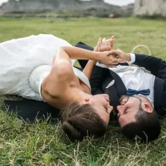 Para młoda leży na trawie podczas sesji plenerowej ślubnej, trzymając się za ręce. W tle ruiny zamku.