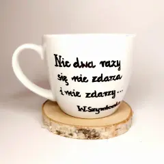 Biały kubek z cytatem Wisławy Szymborskiej "Nic dwa razy się nie zdarza..." na drewnianym podkładzie.