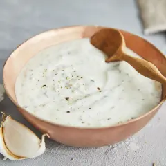 Ein cremiger Knoblauch Joghurt Dip in einer Kupferschale, garniert mit Pfeffer und Kräutern. Daneben liegen frische Knoblauchzehen.