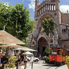 En charmig spårvagn passerar framför en kyrka i Soller, Palma. Folk sitter vid uteserveringar under parasoll.