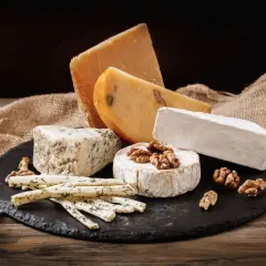 Selezione di formaggi grassi: un pezzo di formaggio erborinato, un brie, un formaggio stagionato e bastoncini di formaggio alle erbe, con noci e tela di juta su un tagliere di ardesia.