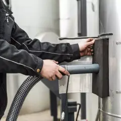 Pracownik czyści komin przez drzwi wyczystki kominowej, używając węża.