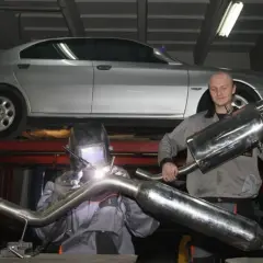 Mechanik w masce spawalniczej wykonuje spawanie wydechu. Obok stoi kolega z gotową częścią.
