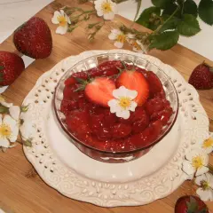 Słodka frużelina truskawkowa w miseczce, ozdobiona połówkami truskawek i białym kwiatkiem. Wokół świeże truskawki i dzikie róże.