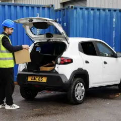 Pracownik w kamizelce odblaskowej wkłada paczkę do bagażnika białego Dacia Spring Cargo, gotowego do dostawy.
