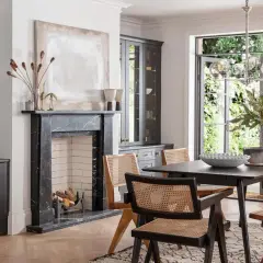 Modern dining room with dark grey cabinets, marble fireplace, and a dark wood table with cane-backed chairs, showcasing current trends in dining room furniture.