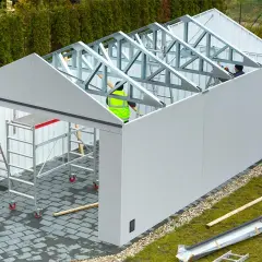 Budowa garażu blaszanego w toku. Zastanawiasz się, ile kosztuje pozwolenie na budowę garażu blaszanego? Na placu budowy widać pracowników i rusztowania.