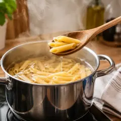 Gotowanie makaronu penne w garnku. Drewniana łyżka z kilkoma rurkami makaronu unosi się nad parującą wodą.