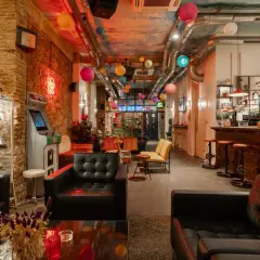 Interno di un social club a Barcellona con divani neri, un cabinato arcade, lanterne colorate e un bancone bar in legno.