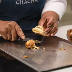 Manos cortan una empanada dorada, revelando su relleno. Otra masa empanada espera en una tabla.
