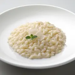 Un cremoso risotto in cagnone, guarnito con parmigiano e una fogliolina verde, servito in un piatto bianco.
