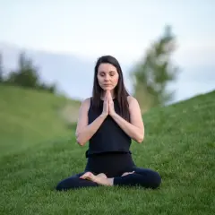 Mujer en posición de loto en la hierba, con las manos juntas en oración. Su rutina de yoga al aire libre es un momento de paz.
