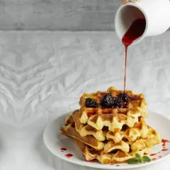 Pila di waffle dorati con more e sciroppo rosso versato da una caraffa. Ecco come fare i waffle perfetti per la colazione.