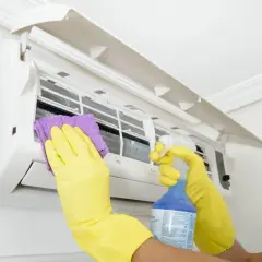 Man con guantes amarillos limpia un aire acondicionado con un paño morado y spray. Aprende como desatascar un aire acondicionado.